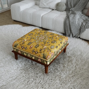 Può includere: Un pouf quadrato caratterizzato da una superficie superiore gialla con un motivo floreale e a tralci multicolore. I lati del pouf sono rivestiti in tessuto verde con un disegno floreale abbinato, sostenuto da quattro gambe corte in legno marrone scuro.