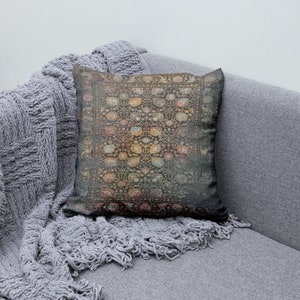 K&ouml;nnte beinhalten: Ein dekoratives quadratisches Kissen mit einem Vintage-Teppichdesign. Das Kissen hat einen dunklen Hintergrund mit einem sich wiederholenden Blumenmuster in Orange-, Gelb- und Blaut&ouml;nen. Es liegt auf einem grauen Sofa mit einem grauen Strickwurf.
