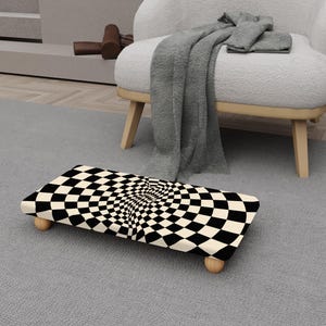 Puede incluir: Un reposapiés rectangular con un diseño de tablero de ajedrez en blanco y negro. La superficie superior crea una ilusión óptica en espiral. Tiene cuatro patas pequeñas de madera redondeadas. El reposapiés está sobre una alfombra gris. Mide unos 60 cm de largo.