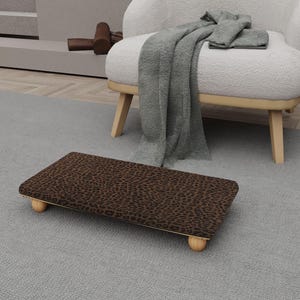 May include: A rectangular footstool with a brown leopard print fabric top and four rounded wooden legs. The footstool is placed on a gray rug, with a white armchair and a gray blanket in the background.