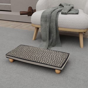 May include: A rectangular footstool with a leopard print top and a black border. The footstool is supported by four small, rounded wooden legs. A gray rug and a white armchair are in the background.