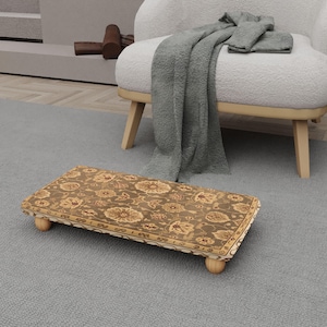 May include: A rectangular footstool with a floral pattern in shades of brown, beige, and gold. The footstool has rounded wooden legs and a decorative border. It is placed on a gray rug near a white armchair.