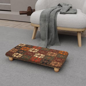 May include: A rectangular footstool with a patterned fabric top in shades of brown, orange, green, and cream. The footstool has four rounded wooden legs. The footstool is placed on a gray carpet.