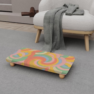 May include: A rectangular footstool with a colorful, retro-style pattern of concentric circles in shades of orange, yellow, pink, and green. The footstool has rounded wooden legs and is placed on a gray carpet.