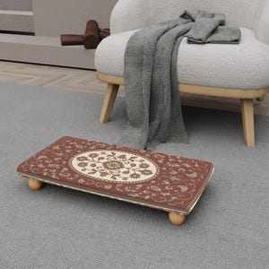 Puede incluir: Un reposapiés rectangular con un diseño floral. El reposapiés tiene un fondo rojo con un centro floral ovalado color crema. Los bordes están adornados en dorado y descansa sobre cuatro pequeñas patas de madera redondeadas.