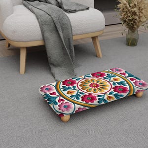 May include: A rectangular footstool with a floral design. The top features a white background with a central yellow and orange floral pattern, surrounded by pink and teal flowers. The stool has four wooden legs and is placed on a gray rug.