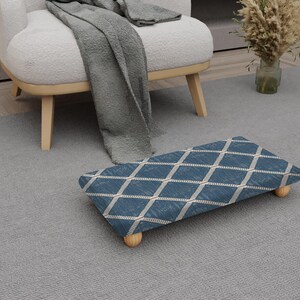 May include: A blue and white patterned footstool with wooden legs. The footstool has a diamond pattern design and is placed on a grey carpet. A white armchair and a grey blanket are in the background.