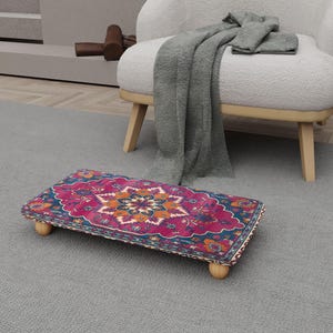 May include: A rectangular footstool with a colorful, patterned top featuring a floral design in shades of pink, blue, orange, and yellow. The footstool is supported by four short, rounded wooden legs, and is placed on a gray rug.