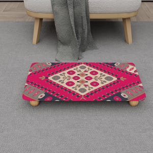 May include: A rectangular footstool with a vibrant pink and cream geometric pattern. The design features a central diamond shape with floral motifs and is supported by four small, light-coloured wooden legs. The footstool is placed on a grey carpet.