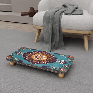 Peut inclure: Un repose-pieds rectangulaire avec un fond turquoise et un motif floral central orange, marron et violet. Le repose-pieds a des pieds en bois arrondis et est posé sur un tapis gris. Le design rappelle un tapis persan.