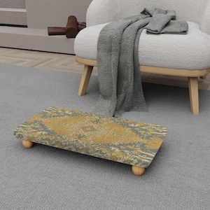 May include: A rectangular footstool with a patterned top in shades of gold and grey, supported by four small, rounded wooden legs. The footstool is placed on a grey carpet, near a white armchair with a grey blanket draped over it.