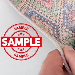 May include: A close-up of a colorful rug with a white border. The rug has a geometric pattern and is labeled "SAMPLE" in a red circle.