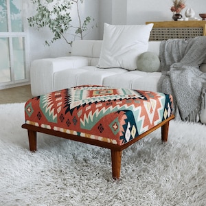 Può includere: Un pouf rettangolare con un motivo geometrico nei toni del rosso, verde acqua e crema. La struttura in legno ha quattro gambe. Il pouf è posto su un soffice tappeto bianco in un salotto.