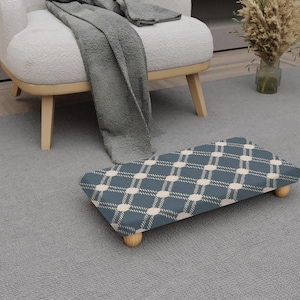 May include: A blue footstool with a cream diamond pattern. The footstool has four wooden legs and is on a grey rug. A grey blanket is draped over a white armchair in the background.