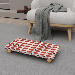 Puede incluir: Un reposapiés rectangular con un patrón retro de óvalos rojos y diamantes azul marino sobre un fondo crema. El reposapiés tiene cuatro patas de madera y está colocado sobre una alfombra gris.