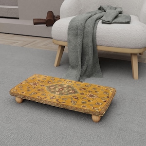May include: A rectangular, yellow footstool with a floral pattern and wooden legs. The footstool is placed on a gray carpet, with a white armchair and a gray blanket in the background. The footstool is approximately 18 inches long.