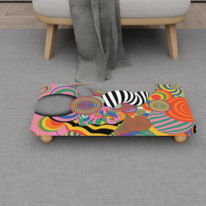 May include: A rectangular footstool with a vibrant, psychedelic pattern. The design features swirling shapes in shades of pink, orange, yellow, green, and black. The footstool has four wooden legs and is placed on a gray carpet.