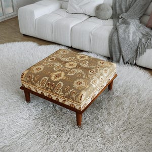 Op de afbeelding: Een vierkante poef met een beige en bruine stoffen bekleding met bloemenpatroon. Het meubelstuk rust op vier korte, donkerbruine houten poten en heeft een decoratieve rand met een herhalend patroon langs de onderkant van het kussen.