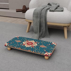 May include: A rectangular footstool with a teal floral pattern and wooden legs. The pattern features red, cream, and gold floral designs. The footstool is placed on a gray carpet near a white armchair.