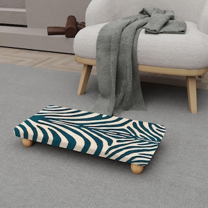 May include: A rectangular footstool with a teal and white zebra stripe pattern. The footstool has four small, rounded wooden legs. The stool is placed on a gray carpet, with a white chair and blanket in the background.
