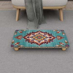 Puede incluir: Un reposapiés rectangular con fondo turquesa y un diseño floral central en tonos naranja, morado y amarillo. El reposapiés tiene patas de madera y un borde estampado. Mide aproximadamente 46 cm de largo y 30 cm de ancho.