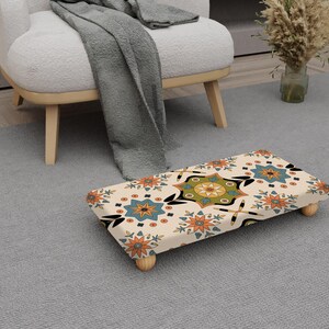 May include: A patterned footstool featuring a cream background with a repeating floral design in orange, blue, and green. The footstool, with four wooden legs, sits on a grey rug. The design is reminiscent of a traditional rug.