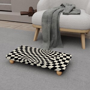 Puede incluir: Una cama rectangular para mascotas con un patrón de tablero de ajedrez en blanco y negro. La cama tiene patas de madera redondeadas y está colocada sobre una alfombra gris. Una manta y una silla grises están en el fondo.