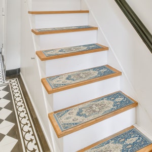 Puede incluir: Escalera con contrahuellas blancas y pelda&ntilde;os de madera. Cada pelda&ntilde;o presenta una alfombra rectangular con un dise&ntilde;o ornamental azul, beige y marr&oacute;n. La escalera tiene una barandilla negra y est&aacute; en un interior bien iluminado.