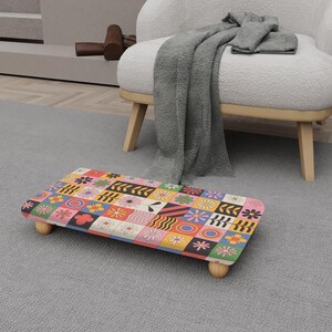 Puede incluir: Un reposapiés rectangular con un colorido diseño de patchwork con flores y formas geométricas. El reposapiés tiene un marco de madera y patas de madera redondeadas. La paleta de colores incluye rosa, naranja, amarillo, negro y verde.