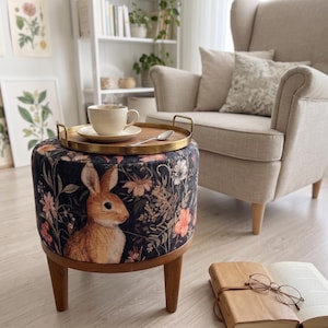 May include: A round ottoman with a rabbit and floral design. The ottoman has a dark background with a brown rabbit and pink and white flowers. A gold tray with a cup and saucer sits on top. The ottoman has wooden legs.