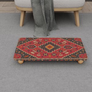 Puede incluir: Un reposapiés rectangular con un diseño estampado en rojo, negro y naranja. El reposapiés tiene un marco de madera y está sostenido por cuatro patas de madera pequeñas y redondeadas. El patrón se asemeja a un diseño de alfombra tradicional.