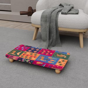 May include: A rectangular footstool with a colorful patchwork design featuring various patterns and images, including tigers and pineapples. The footstool has a wooden frame and four rounded wooden legs. The color palette includes pink, blue, orange, and green.