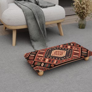 May include: A rectangular footstool with a patterned rug design in red, black, and beige. The footstool has four wooden legs and is on a grey carpet. The design features geometric shapes and traditional motifs.