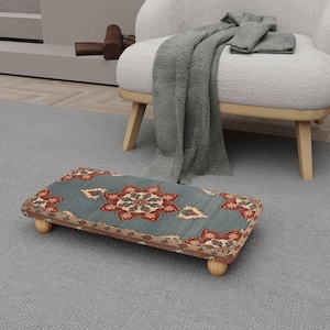 Op de afbeelding: Een rechthoekige voetenbank met een teal en rood bloemenpatroon. De voetenbank heeft een houten frame en vier afgeronde houten poten. Het ontwerp doet denken aan een traditioneel tapijt.