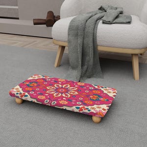 May include: A rectangular footstool with a vibrant floral pattern in shades of pink, orange, and blue. The stool has a wooden base and four small, rounded wooden legs. The design is reminiscent of a traditional rug.