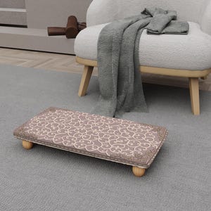 May include: A rectangular footstool with a patterned top and wooden legs. The top has a repeating swirl design in beige and mauve, framed by a darker border. The legs are short, rounded, and made of light-coloured wood.