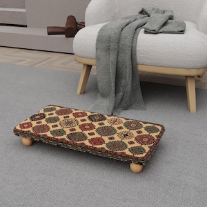 May include: A rectangular footstool with a patterned top in shades of red, green, and brown, resting on four small, rounded wooden legs. The footstool is placed on a gray carpet, with a white armchair and a gray blanket in the background.