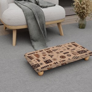 May include: A rectangular footstool with a beige background and a repeating pattern of brown geometric shapes. The footstool has four small, rounded wooden legs and is placed on a grey rug.