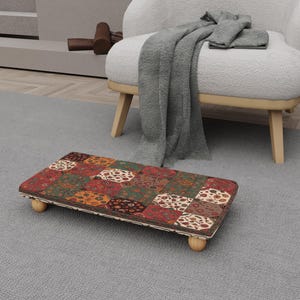 Könnte beinhalten: Ein rechteckiger Fußhocker mit einem Patchwork-Design in Rot-, Grün- und Brauntönen. Der Fußhocker hat einen Holzrahmen und steht auf vier kleinen, abgerundeten Holzbeinen. Das Patchwork-Muster weist verschiedene geometrische und florale Motive auf.