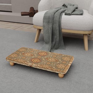 Op de afbeelding: Een rechthoekige voetenbank met een stoffen bovenkant met patronen in bruin, beige en roodtinten. De voetenbank heeft afgeronde houten poten en een decoratieve rand. De voetenbank staat op een grijs tapijt, in de buurt van een witte fauteuil.