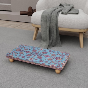Può includere: Un poggiapiedi rettangolare con un motivo floreale nei toni del blu, rosso e oro. Il poggiapiedi ha gambe in legno arrotondate e un piano in tessuto fantasia. Il poggiapiedi è posizionato su un tappeto grigio.