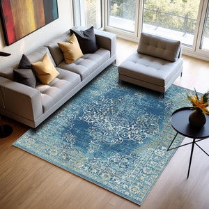 Puede incluir: Una alfombra de salón azul y crema con un diseño vintage desgastado. La alfombra está colocada sobre un suelo de madera en un salón con un sofá gris y un sillón gris.