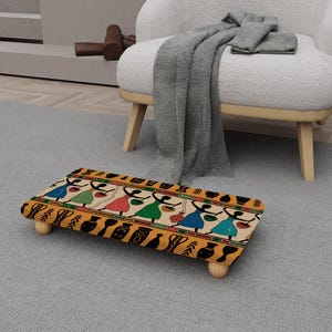 Peut inclure: Un repose-pieds rectangulaire avec un motif coloré représentant des personnages en robes portant des paniers. Le repose-pieds a un fond jaune avec des éléments décoratifs noirs et des pieds en bois.