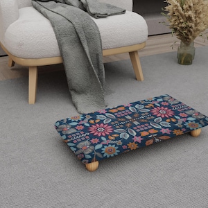 May include: A navy blue footstool featuring a floral design in pink, orange, and blue. The footstool has four short, light-coloured wooden legs. It is placed on a grey carpet. The footstool is approximately 40 cm long.