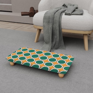 Puede incluir: Un reposapiés rectangular con un diseño retro. La parte superior presenta un patrón de círculos verdes sobre un fondo naranja, delineados en blanco. El taburete tiene cuatro patas cortas de madera de color claro.