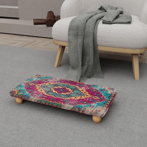May include: A rectangular footstool with a vibrant, patterned design in shades of pink, teal, and gold. The footstool rests on four small, rounded wooden legs, offering a touch of elegance. The design is reminiscent of a traditional rug.