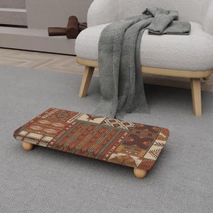 May include: A rectangular footstool with a patterned design in shades of brown, beige, and rust. The footstool has rounded wooden legs and is placed on a grey rug. The design features geometric and tribal-inspired motifs.