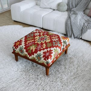 Op de afbeelding: Een vierkante poef met een levendig geometrisch kilimpatroon in rood, crème, groen en roze. Het meubelstuk rust op vier korte, taps toelopende houten poten met een natuurlijke bruine afwerking, wat een stabiele basis vormt voor het zitkussen.