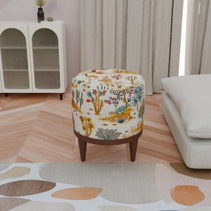 Op de afbeelding: Een ronde, witte poef met een kleurrijke bloemenprint met woestijnplanten en -dieren. De poef heeft houten poten.