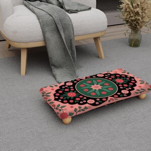 May include: A decorative footstool with a floral design. The footstool has a pink background with a black border and a central green circle. Red and pink flowers and green leaves are scattered across the surface. The footstool is supported by four wooden legs.